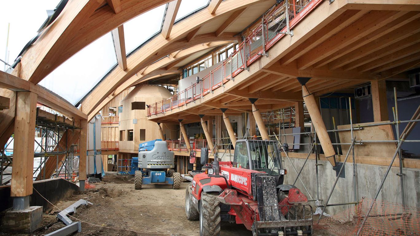 The partially-completed winter garden inside Essex Business School. The ground remains muddy and large construction vehicles are parked where plants will later grow. The woodwork is covered by scaffolding.